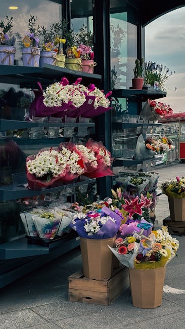 A display of various floral arrangements and bouquets at a flower shop, featuring vibrant white, pink, and purple flowers including gerberas, lilies, and chrysanthemums. The flowers are wrapped in col