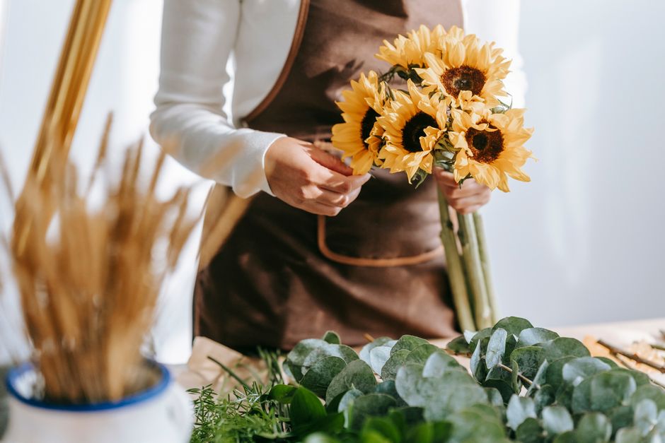 A florist, wearing a white long-sleeved shirt and a brown apron, is holding a bouquet of fresh yellow sunflowers with dark centers and light green stems. The arrangement is held in front of the floris