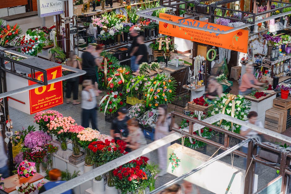 A busy indoor flower market with floral arrangements and bouquets on display, including vibrant pink, red, purple, and white chrysanthemums, roses, and mixed flower displays. The flowers are arranged 