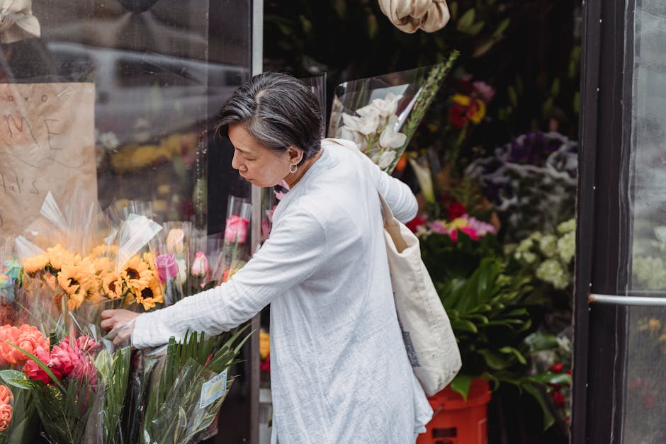 A woman dressed in a white coat selecting vibrant flower bouquets at a flower shop, with arrangements of yellow, pink, and orange flowers wrapped in clear plastic. The background features additional f